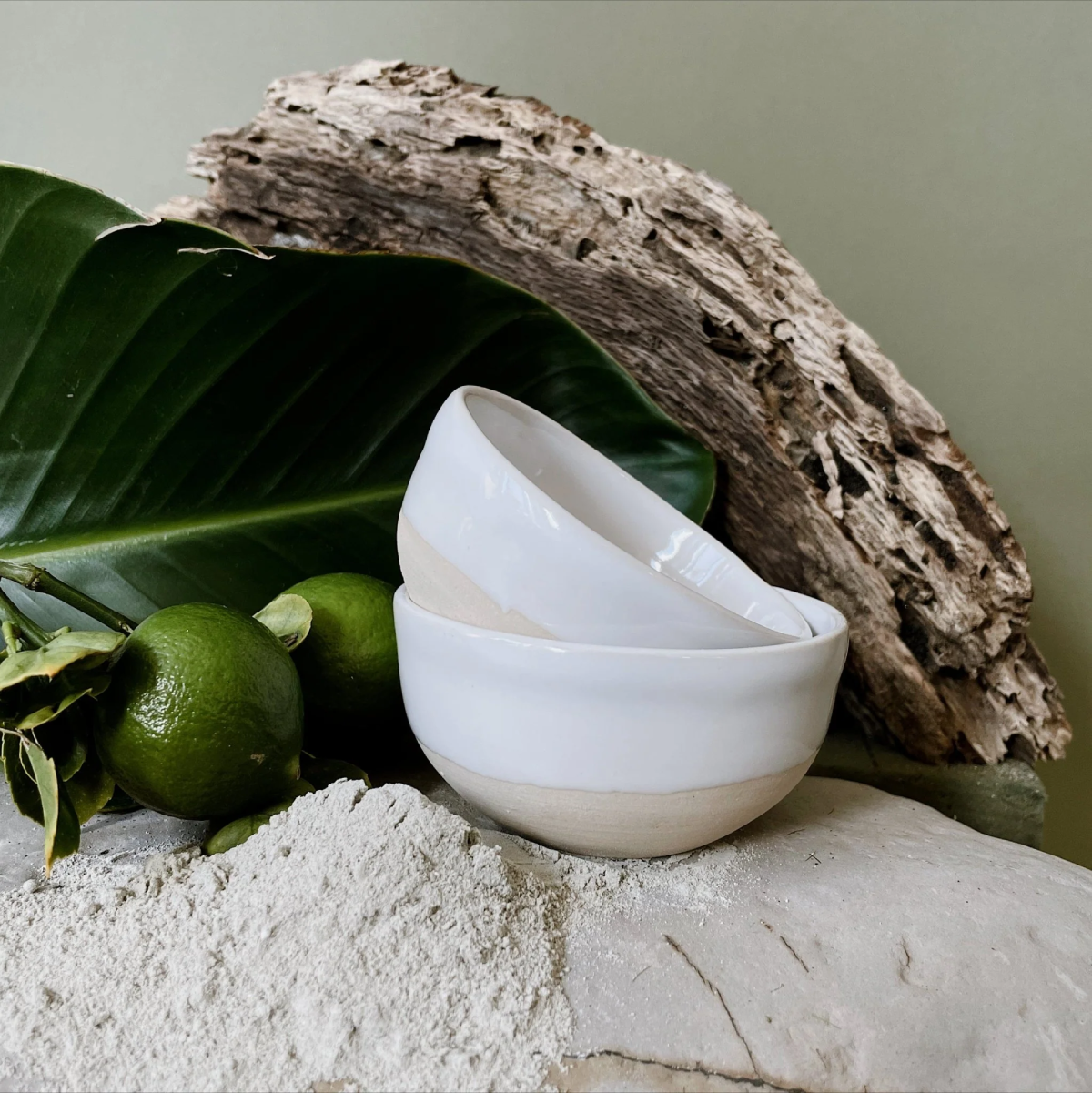 Stack of white ceramic bowls on a stone surface with green leaves and driftwood in the background
